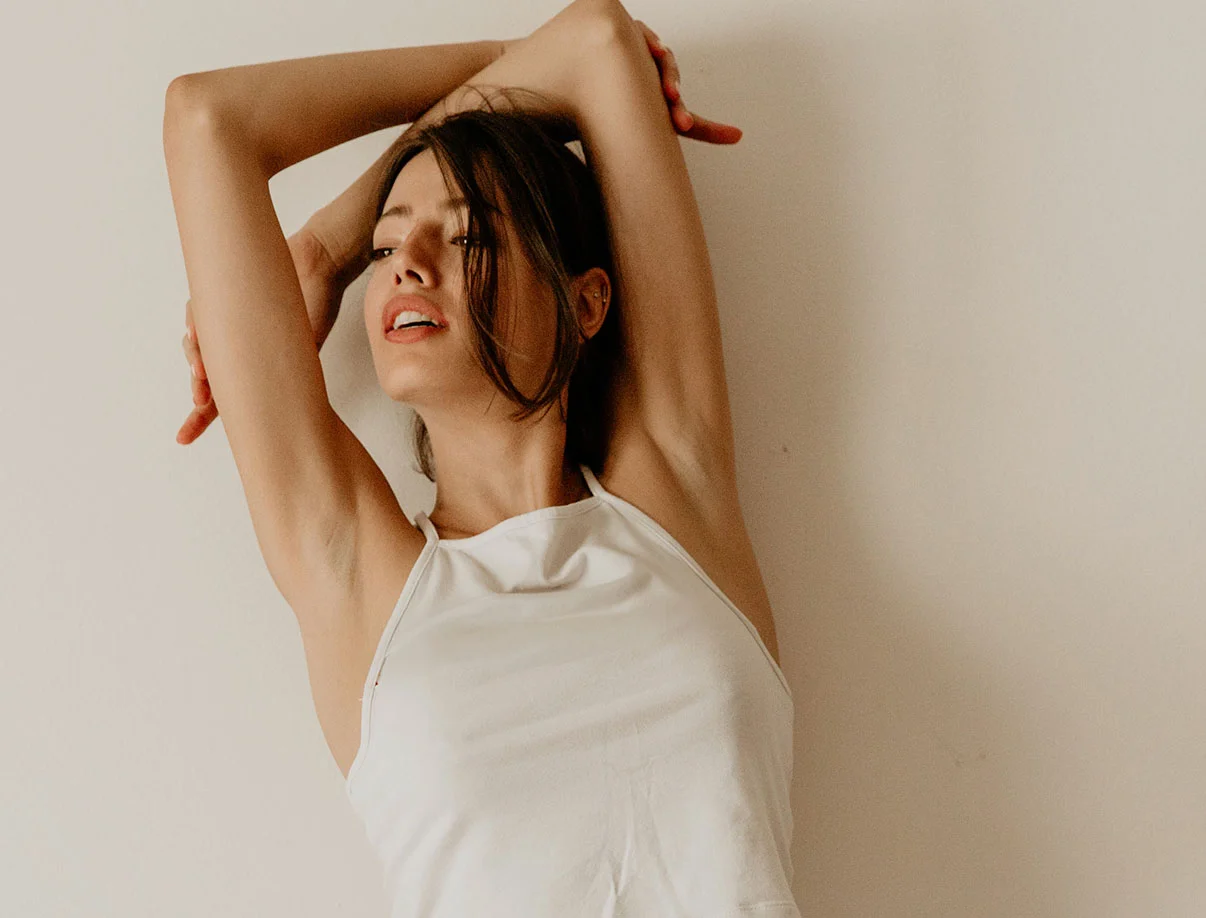 A woman in a white top strikes a relaxed pose, leaning slightly with her arms raised - Arm Lift in West Hollywood, CA