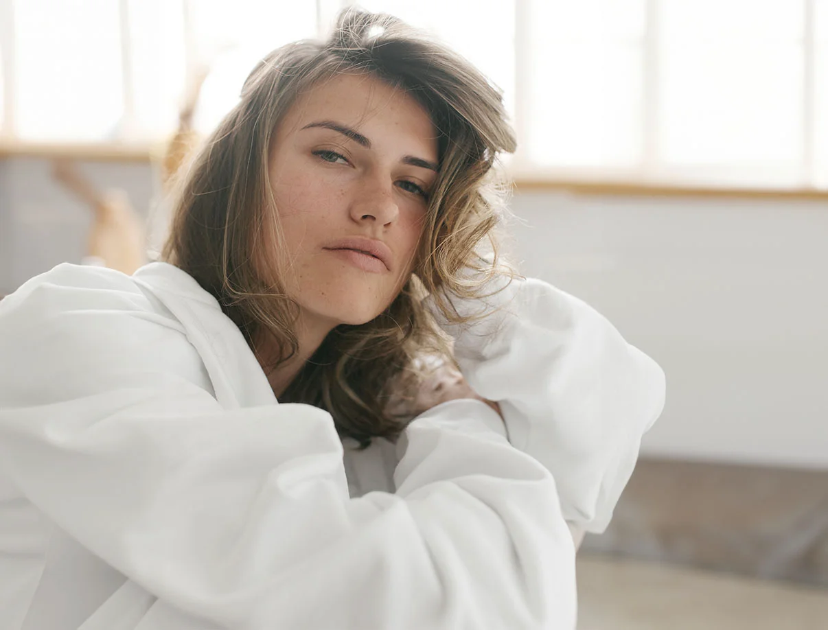A woman with light brown hair, in a white jacket, sitting in a bright room - Brow Lift in West Hollywood, CA
