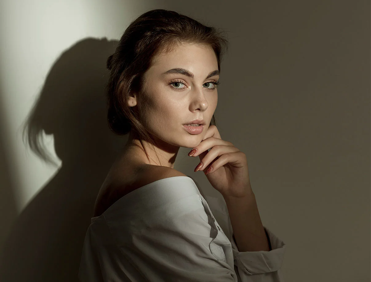 A poised woman in soft lighting, wearing an off-shoulder white top, her hair styled loosely, and gazing thoughtfully - Buccal Fat Removal in West Hollywood, CA