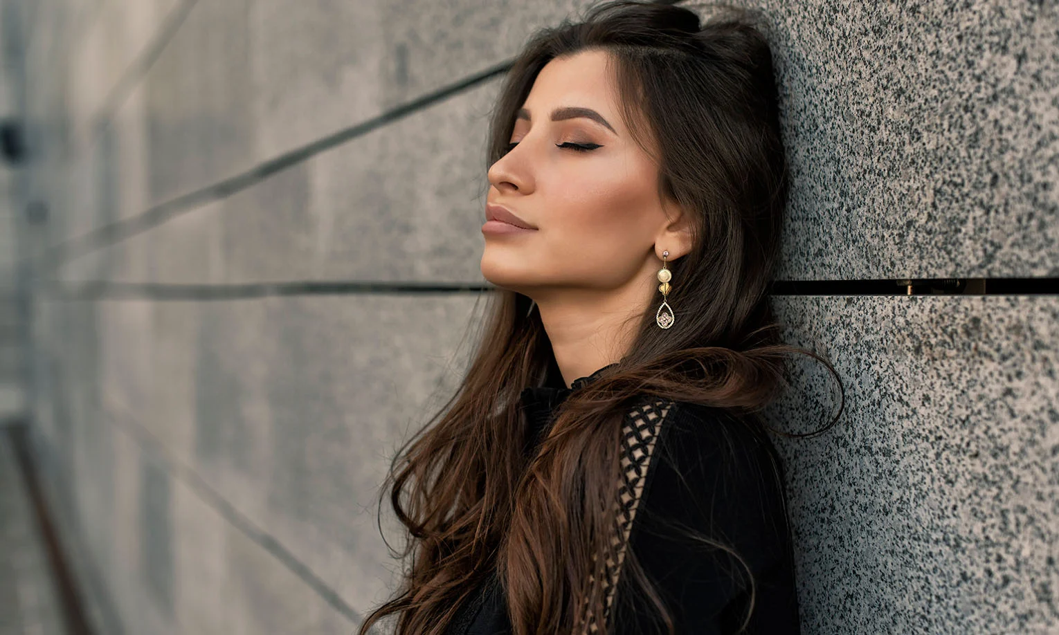 A confident brunette woman leaning against a stone wall, eyes closed, with elegant gold earrings and flowing hair - Buccal Fat Removal in West Hollywood, CA