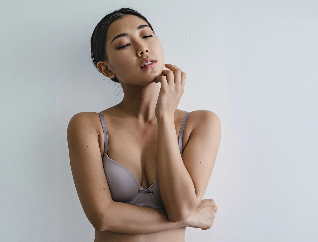 Woman in a gray bra standing against a light-colored wall with her eyes closed, touching her chin - Fat Transfer Breast Augmentation in West Hollywood, CA