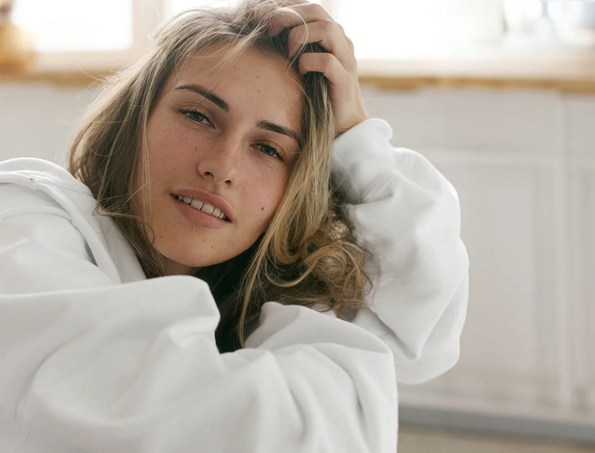 Woman in a white hoodie leaning back and touching her hair, with natural light in the background - FUE Brow Transplant in West Hollywood, CA