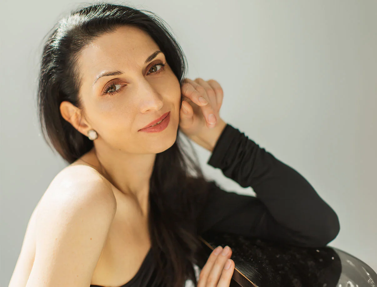 Woman with long dark hair and a black off-shoulder top, smiling gently with her hand resting on her cheek - Lower Eyelid Surgery in West Hollywood, CA