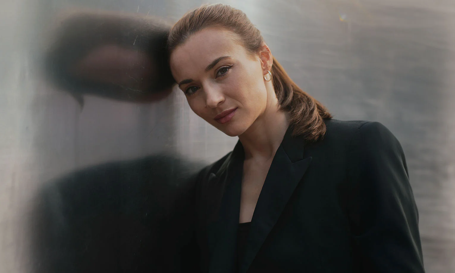 Woman in a black blazer leaning her head against a reflective metallic wall, looking directly at the camera - Lower Eyelid Surgery in West Hollywood, CA