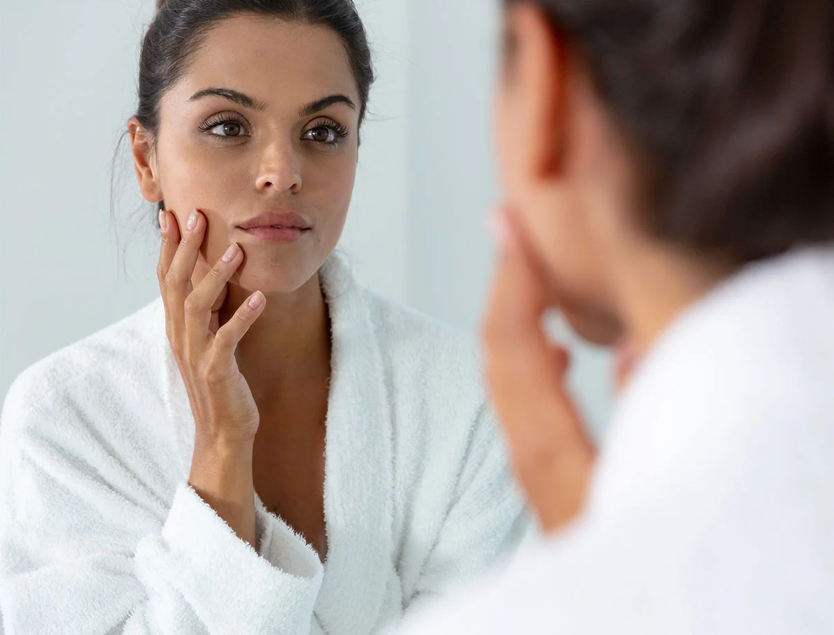 A woman in a white robe examines her reflection in a mirror, thoughtfully touching her face with her hand. - Morpheus 8 in West Hollywood, CA