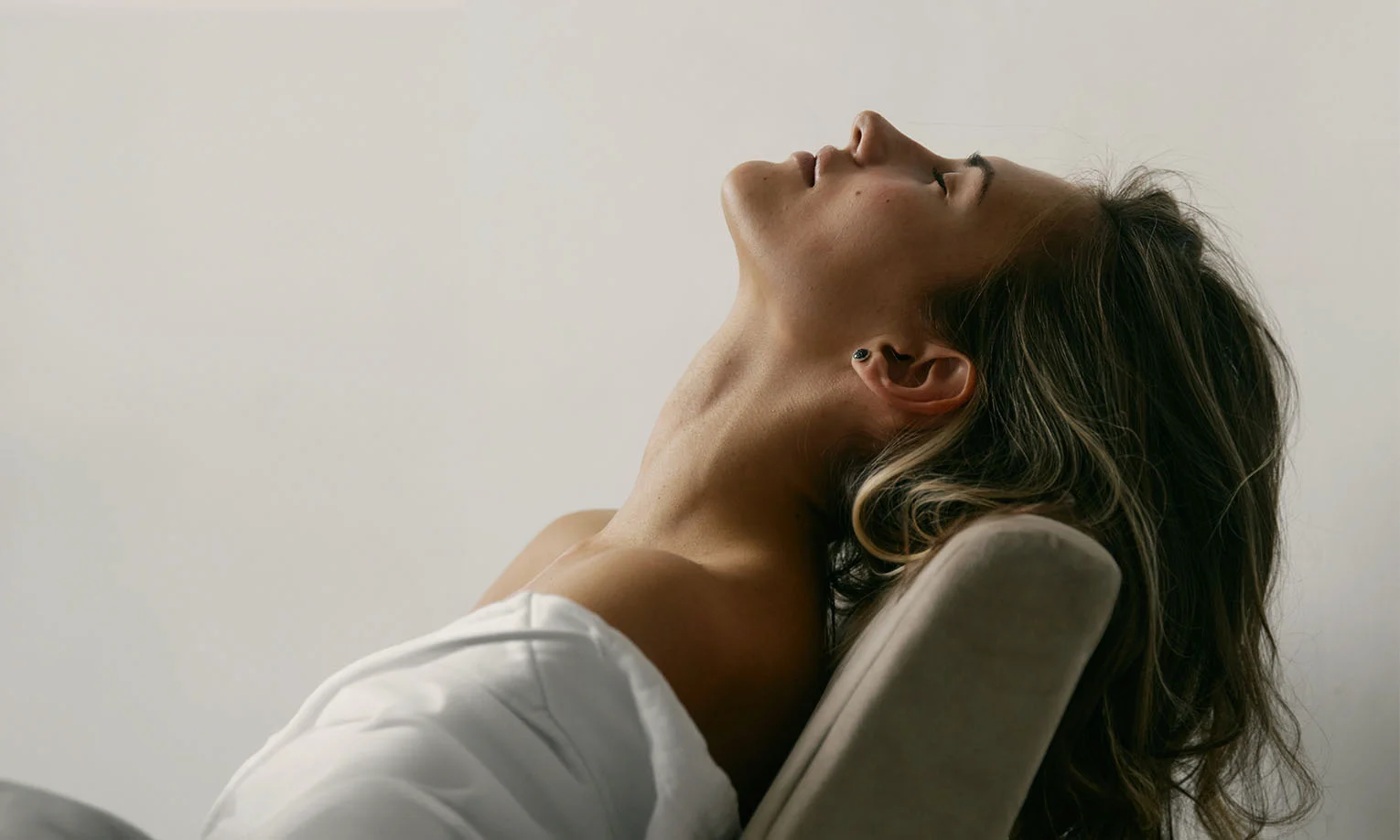 Close-up of a woman leaning back in a chair with her head tilted back, emphasizing her neck and jawline against a light background - Neck Lift in West Hollywood, CA