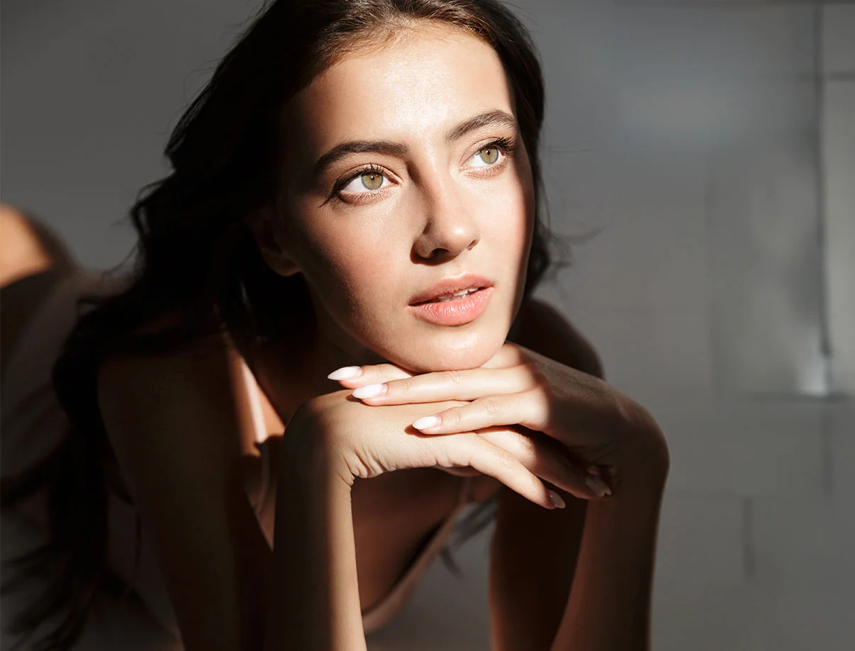 A woman with long dark hair rests her chin on her hands, gazing thoughtfully, with soft light illuminating her features. - Radiesse in West Hollywood, CA
