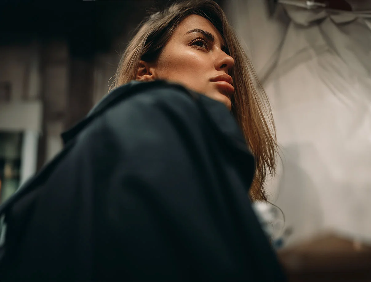 A close-up view of a woman looking upwards, with long hair and wearing a dark coat, against a blurred background. - Sculptra in West Hollywood, CA