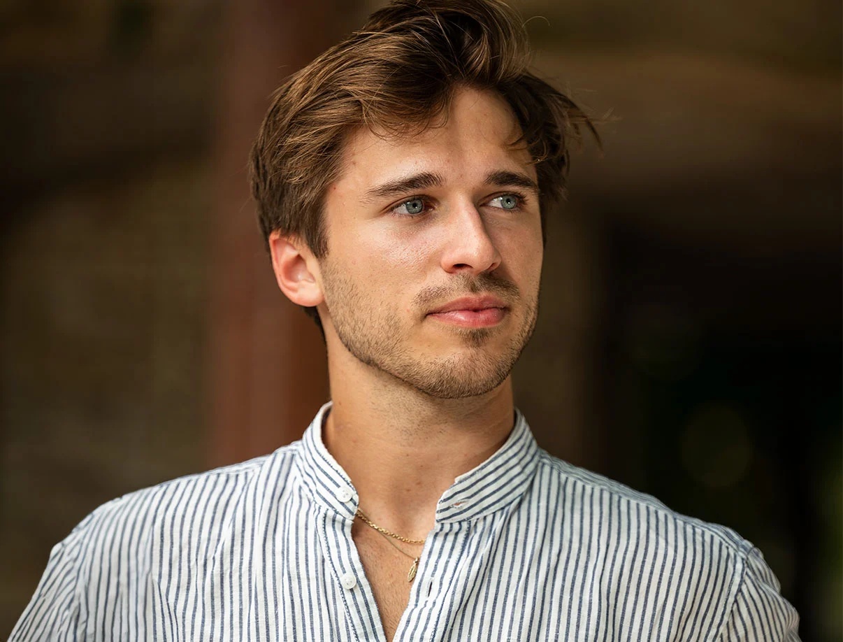 A young man with tousled hair wearing a striped shirt, gazing into the distance, suggesting thoughtfulness and natural elegance - Stelar Hair in West Hollywood, CA