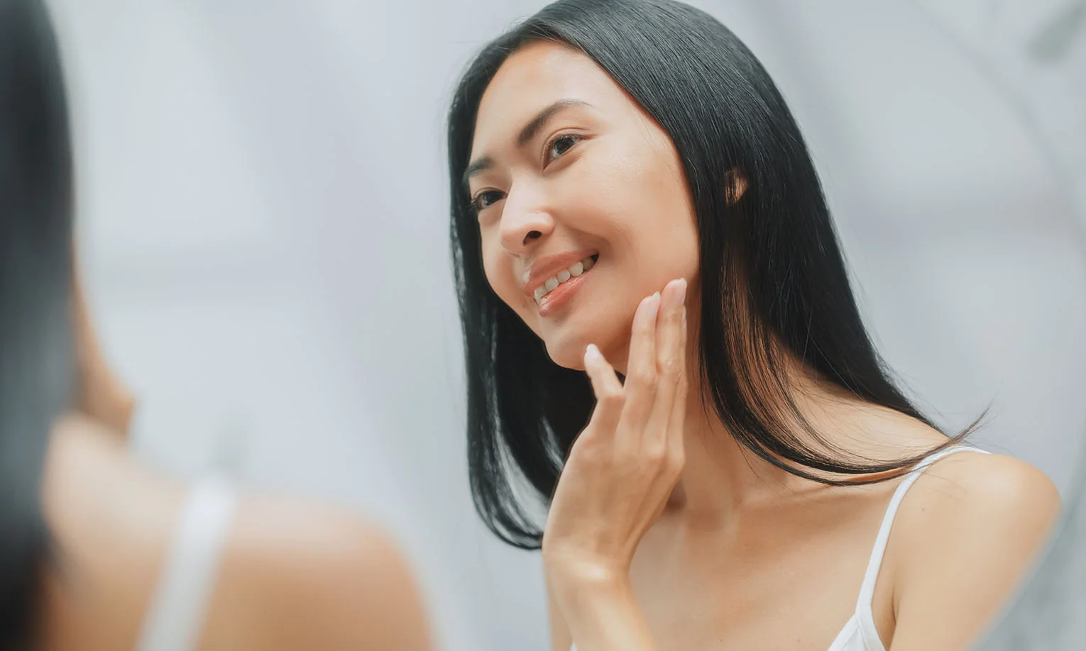 Smiling woman with smooth, straight black hair, gently touching her cheek while looking into a mirror - Submental Liposuction in West Hollywood, CA