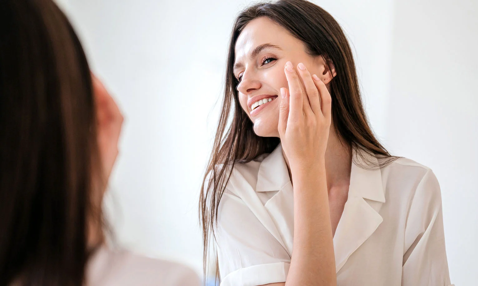 A young woman smiles while applying skincare, looking into a mirror with soft lighting in a bright, minimalistic space. - Sun and Brown Spot Removal in West Hollywood, CA