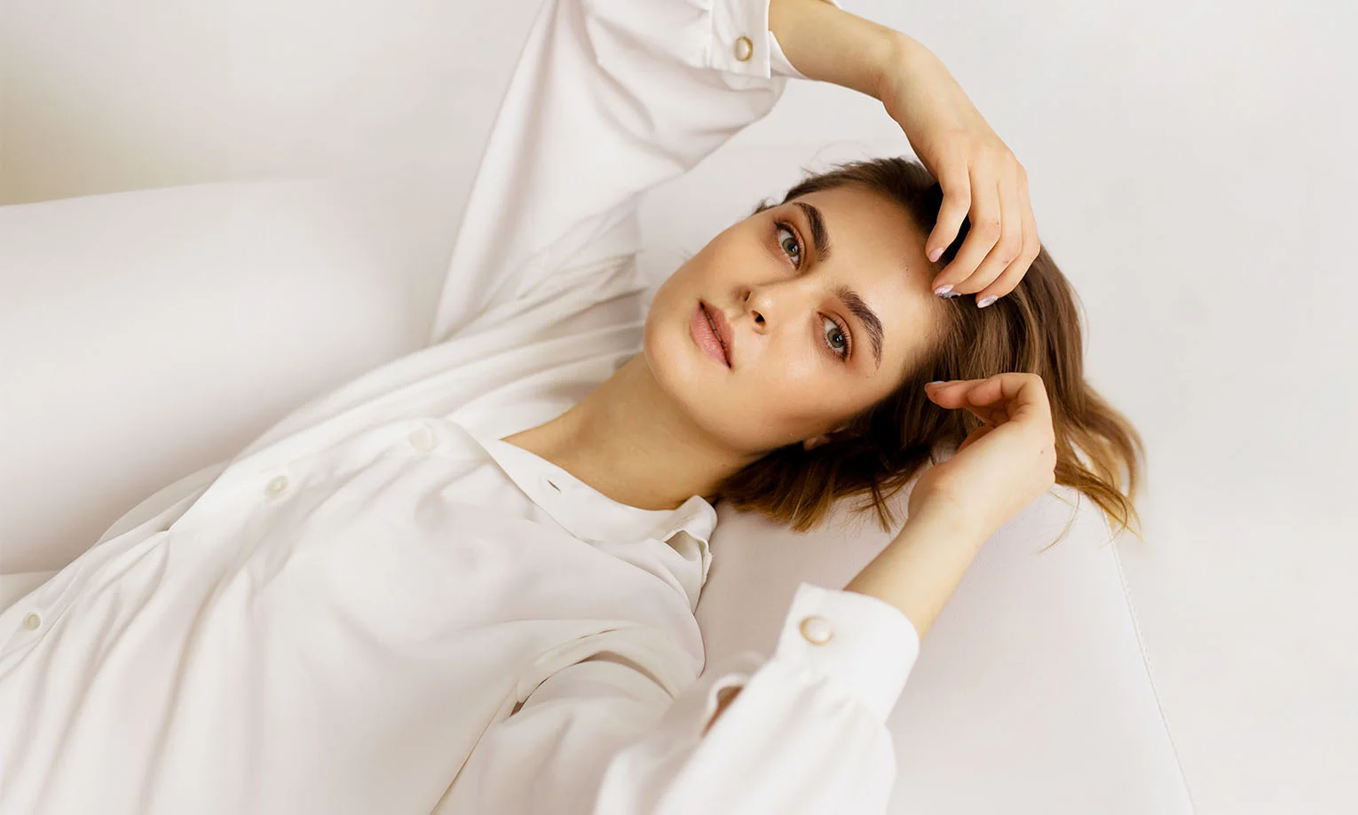 A woman with medium-length hair lies on a white surface, wearing a white button-up shirt and gazing thoughtfully at the camera. - Thread Lifts in West Hollywood, CA
