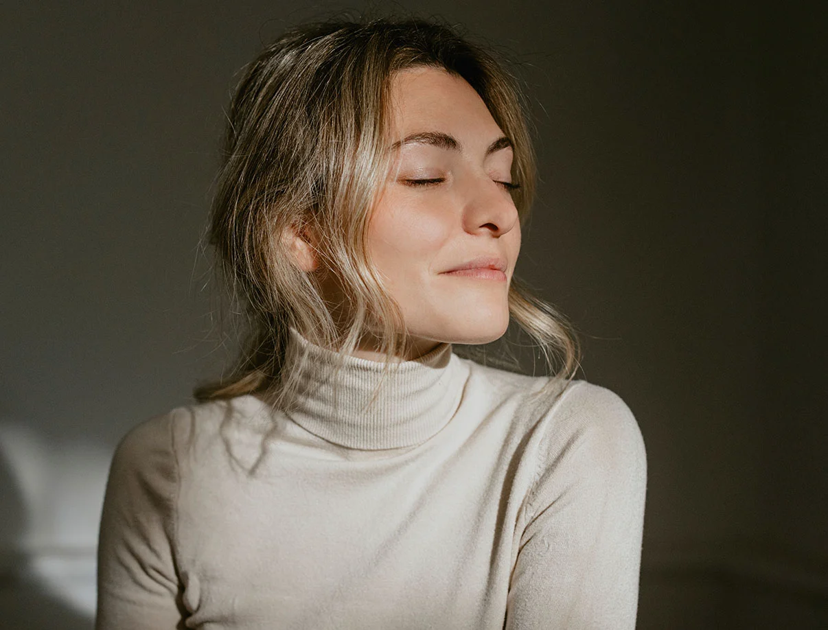 Blonde woman in a cream turtleneck sweater, softly smiling with her eyes closed, enjoying serene natural light - Upper Eyelid Surgery in West Hollywood, CA